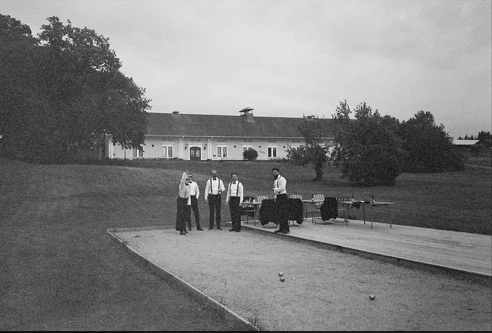 Män i hängslen spelar boule framför herrgården, i svartvitt